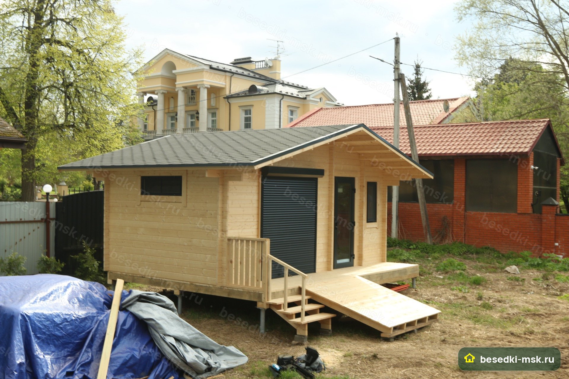 Хозблок фронт 3/4: серая черепица, светлый мини-брус, тёмные рольставни, остеклённая дверь, окно, настил с лесенкой и пандусом